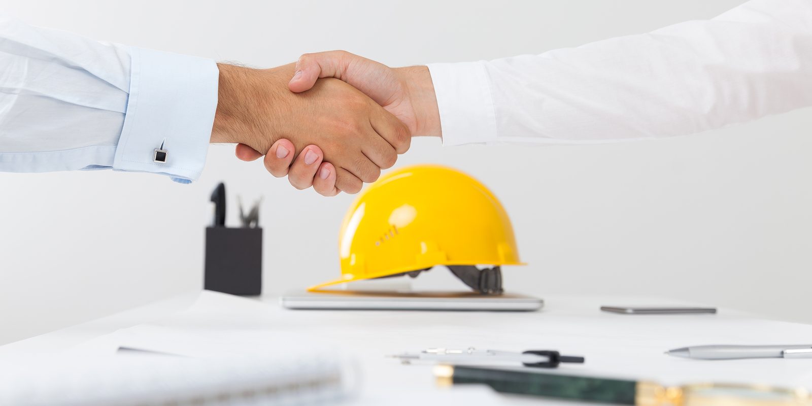 Things to Include in Your Construction Insurance Close up of a handshake and a yellow builder helmet is in the background.