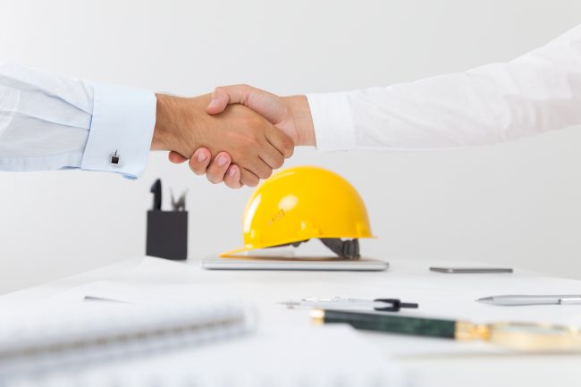 Close up of a handshake and a yellow builder helmet is in the background.